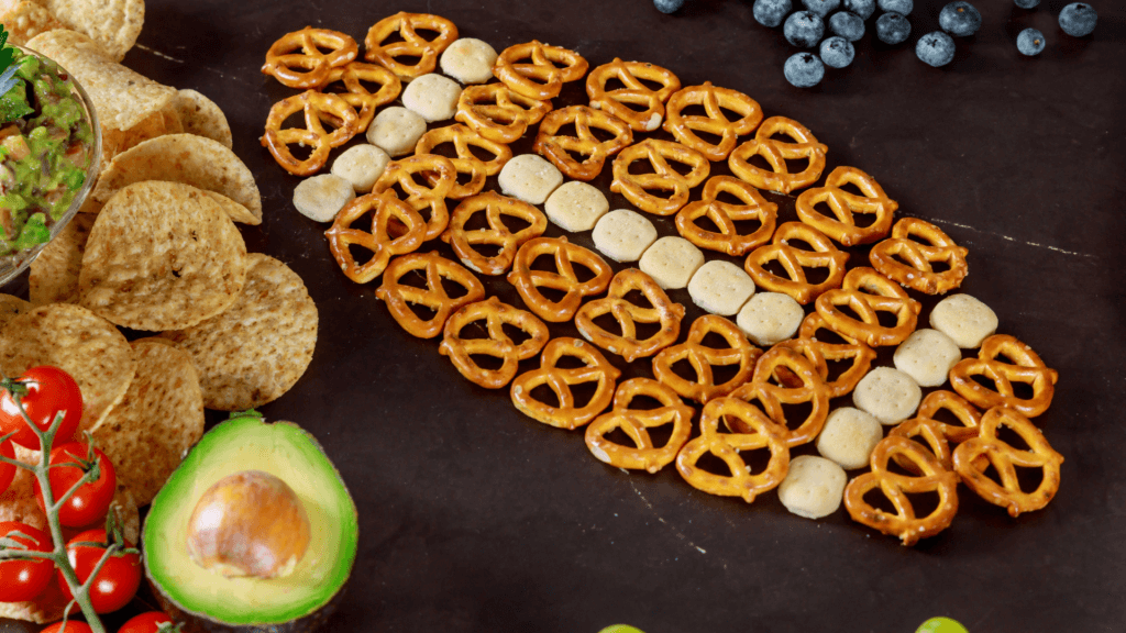 Pretzels and round crackers arranged in a football shape on a dark surface, surrounded by chips, guacamole, avocado, cherry tomatoes, blueberries, and grapes—perfect for your next Indie Big Game Ad party spread.
