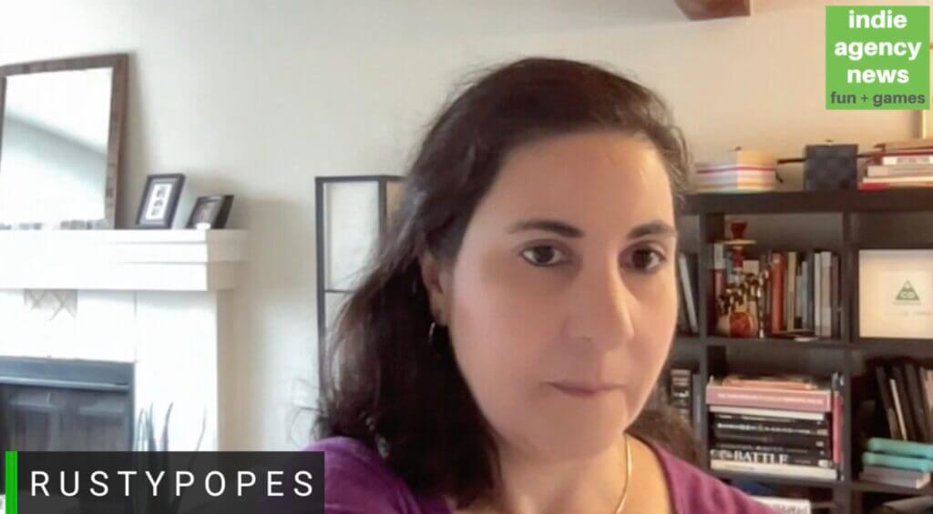 A woman sits indoors with bookshelves and a fireplace in the background. The screen displays text: indie agency news fun + games, RUSTYPOPES, and a shoutout to their latest ‘Pun Fight’ event.