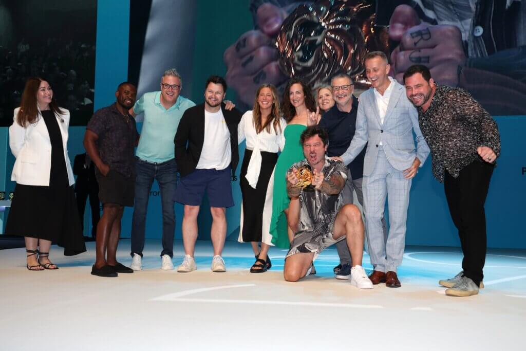 A group of ten people pose on stage at Cannes, one person kneeling and holding a gold lion trophy, all smiling for a celebratory photo against a blue backdrop showcasing standout indie entries.