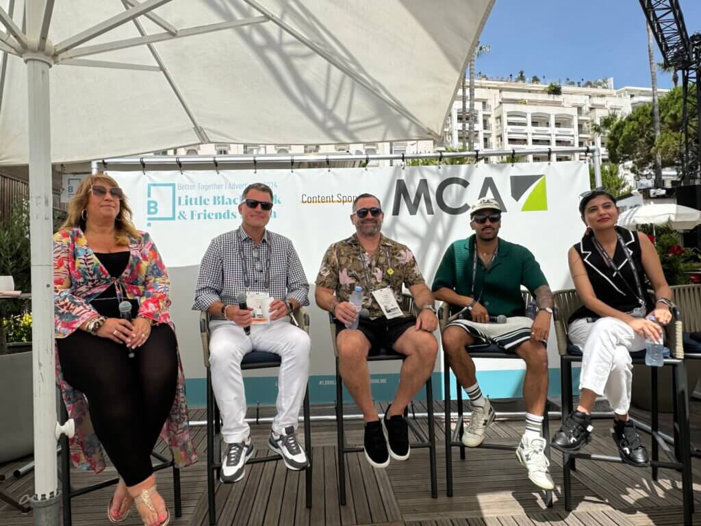 Five people sit on high chairs outdoors in front of a panel banner, posing for a group photo at an industry event, capturing the spirit of indie candor.