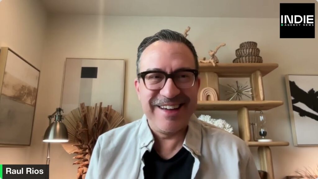 A man with glasses and a mustache smiles while seated indoors, shelves and framed art behind him. The text INDIE AGENCY NEWS appears in the upper right, highlighting marketing trends and audience segmentation.