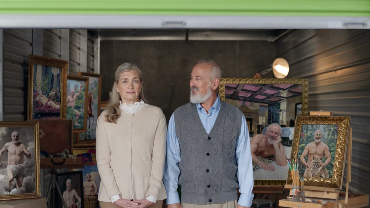An older couple stands in a storage unit filled with framed paintings of a bearded man posing in various humorous, semi-nude settings.