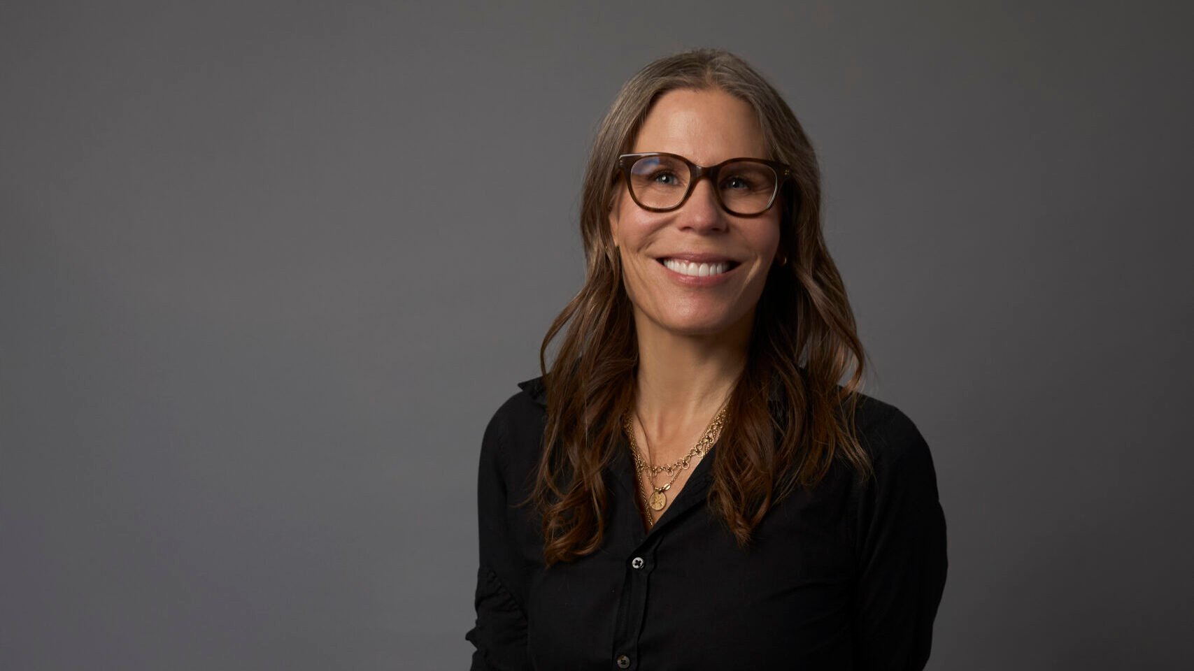 Leslie Barrett, with long brown hair and glasses, wearing a black shirt and pants, sits on a light green stool against a plain gray background.