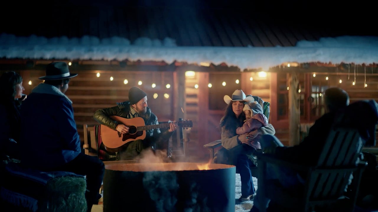 Five people sit around a fire pit at night; one plays a guitar while another holds a child. Lights and a log cabin are visible in the background.
