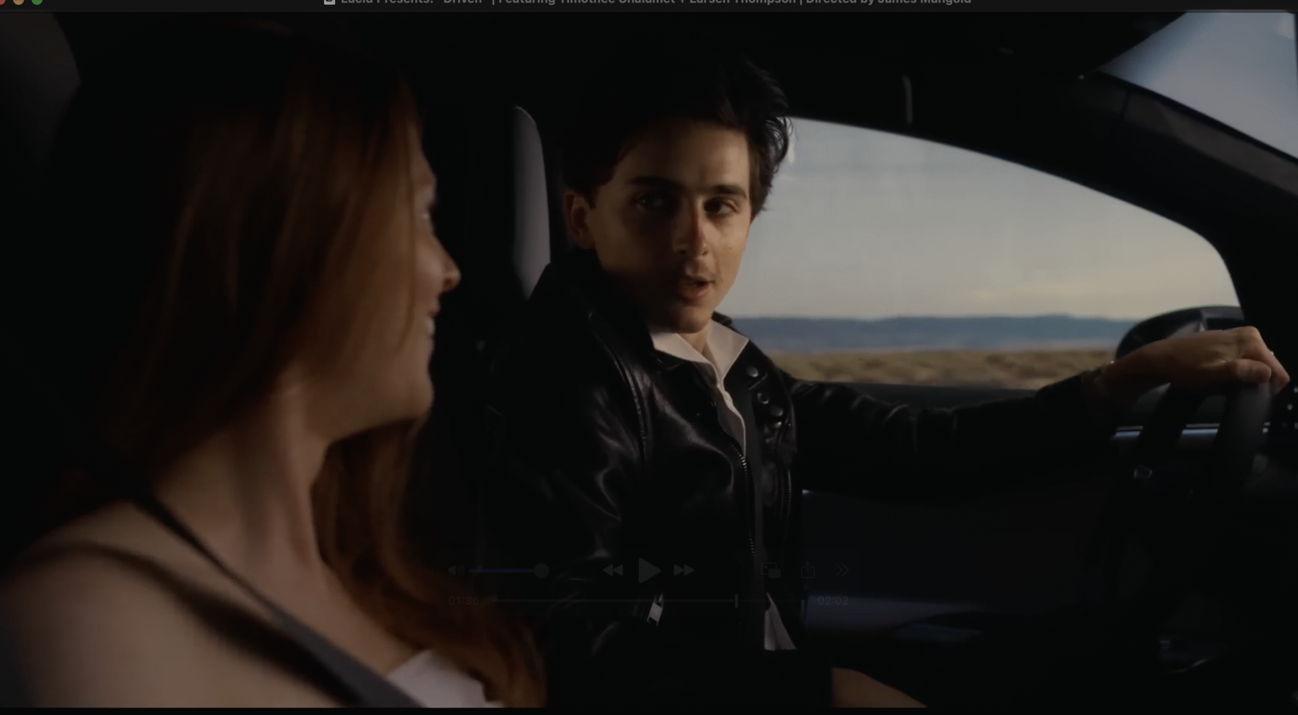 A man driving a Lucid car looks at the woman in the passenger seat while talking; landscape and sky are visible through the window, evoking a cinematic moment reminiscent of a Timothée Chalamet advertising scene.