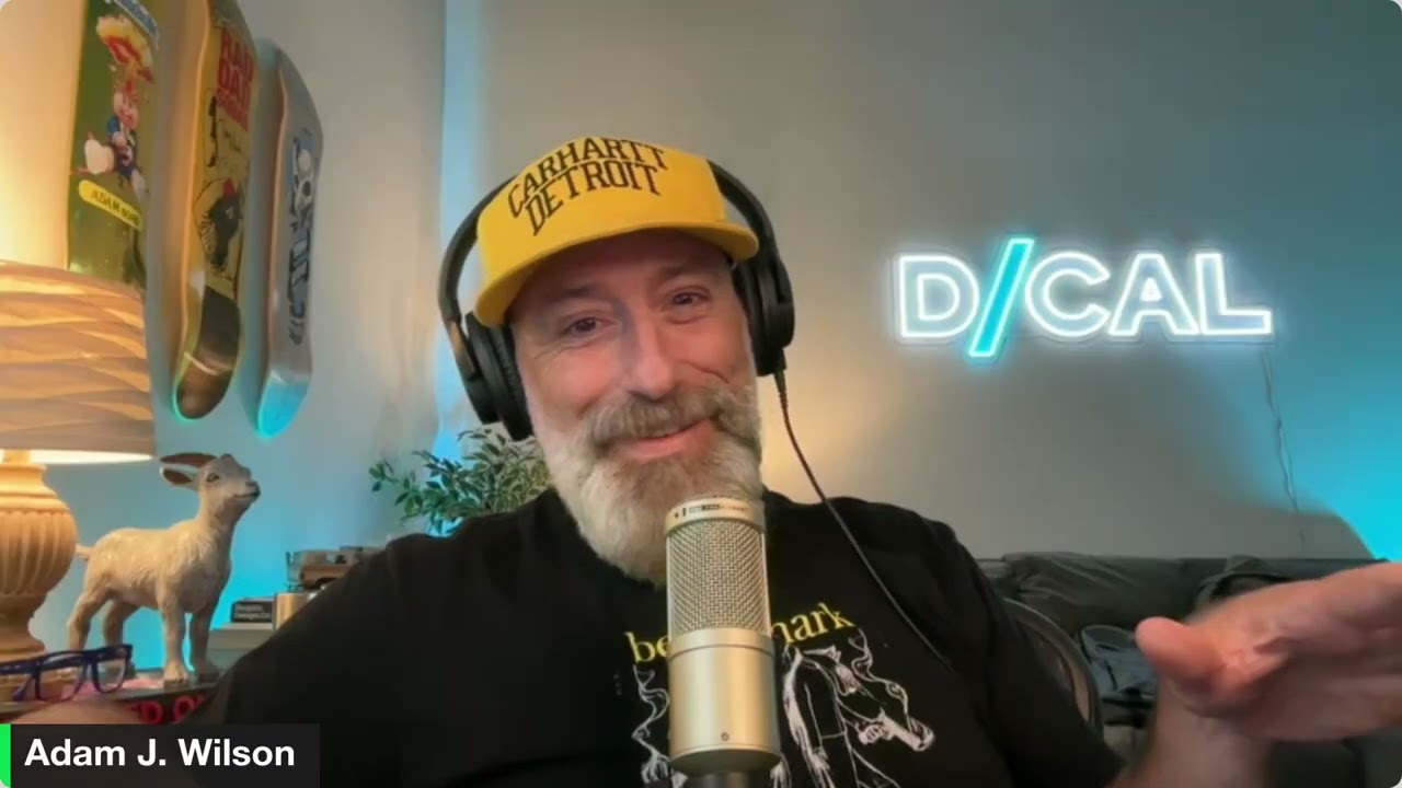 A man with a beard and yellow Carhartt Detroit hat speaks into a microphone, sharing data-driven conversations in a room with a neon D/CAL sign and skateboard decks on the wall.