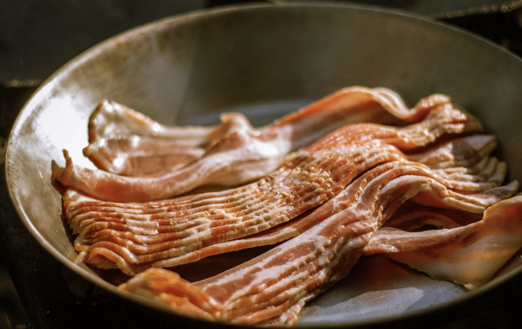 Several raw strips of bacon are laid out in a frying pan, beginning to cook and filling the kitchen with sizzling realness.