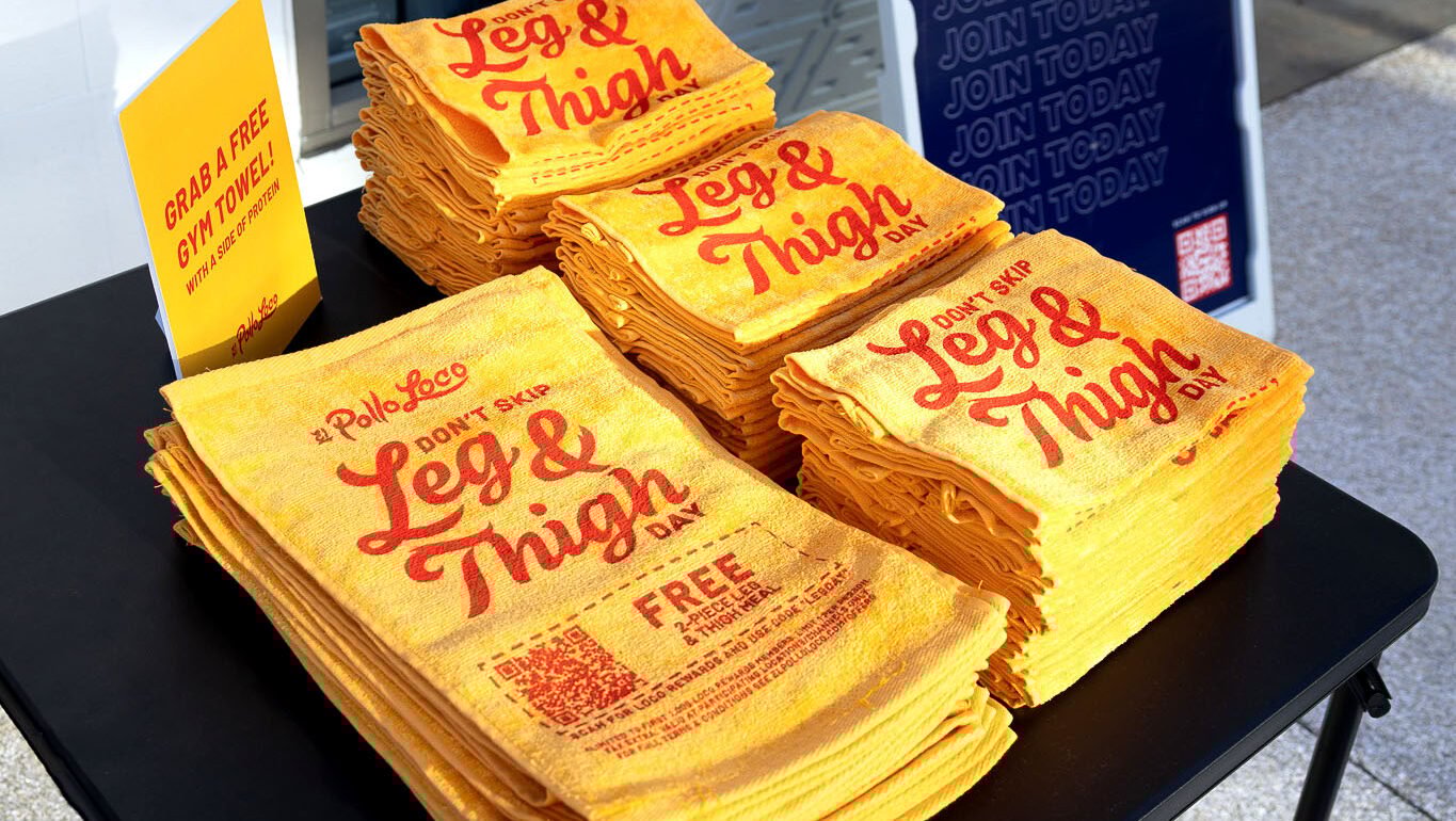 Stacks of yellow towels with bold red Leg & Thigh Day text are displayed on a black table next to a sign that reads Grab a Free Gift Towel from El Pollo Loco.