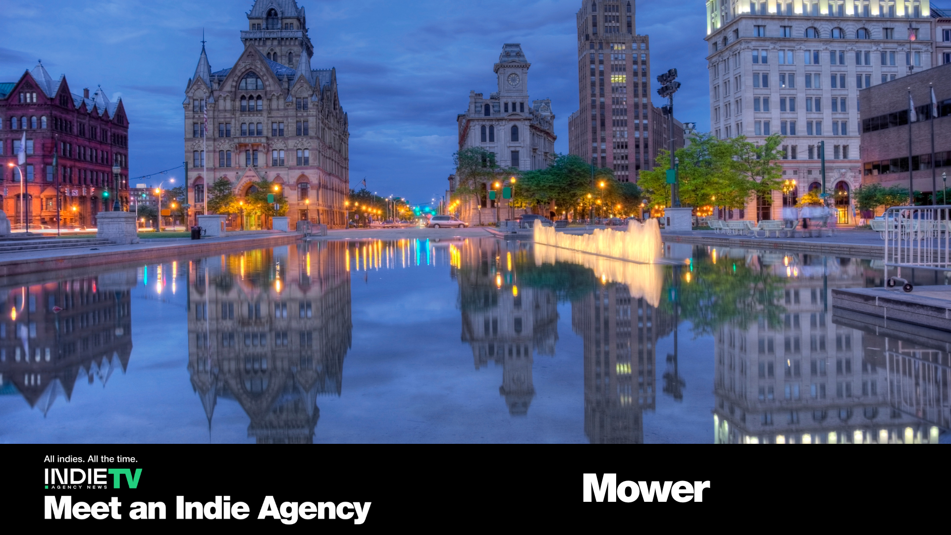 A cityscape at dusk with historic buildings reflected in a calm water feature; text at the bottom reads “Meet an Indie Agency” and “Mower”—where underdogs become friends and bold ideas light up the night.