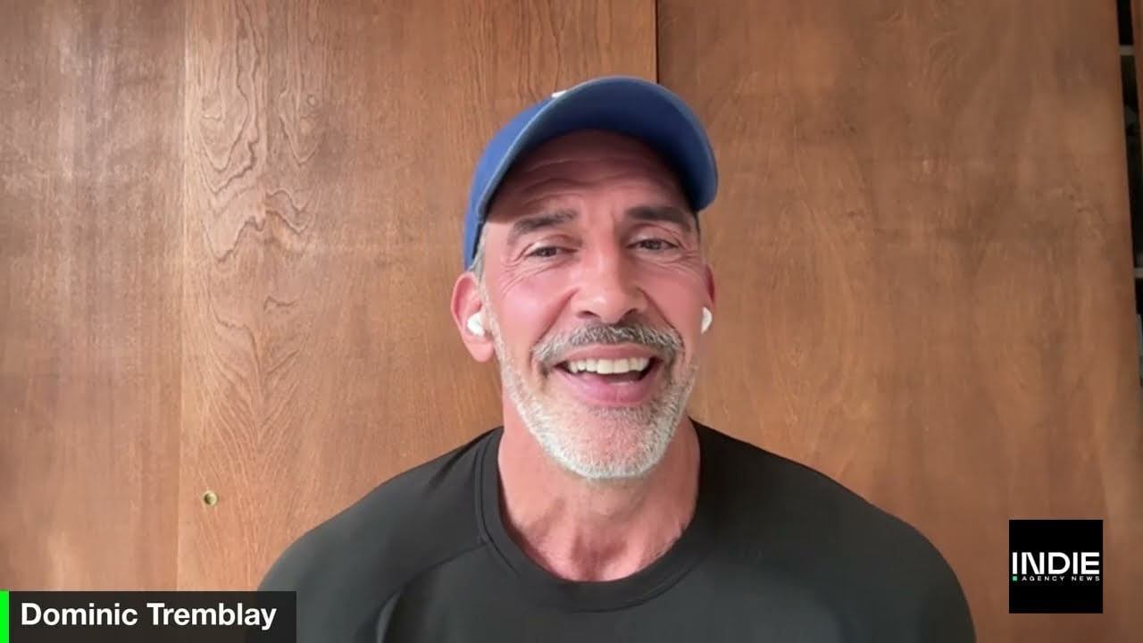 A man with a gray beard wearing a blue cap and black shirt smiles at the camera while using wireless earbuds. Text on screen reads Dominic Tremblay and INDIE, highlighting non-agency people shaping beautiful agencies.
