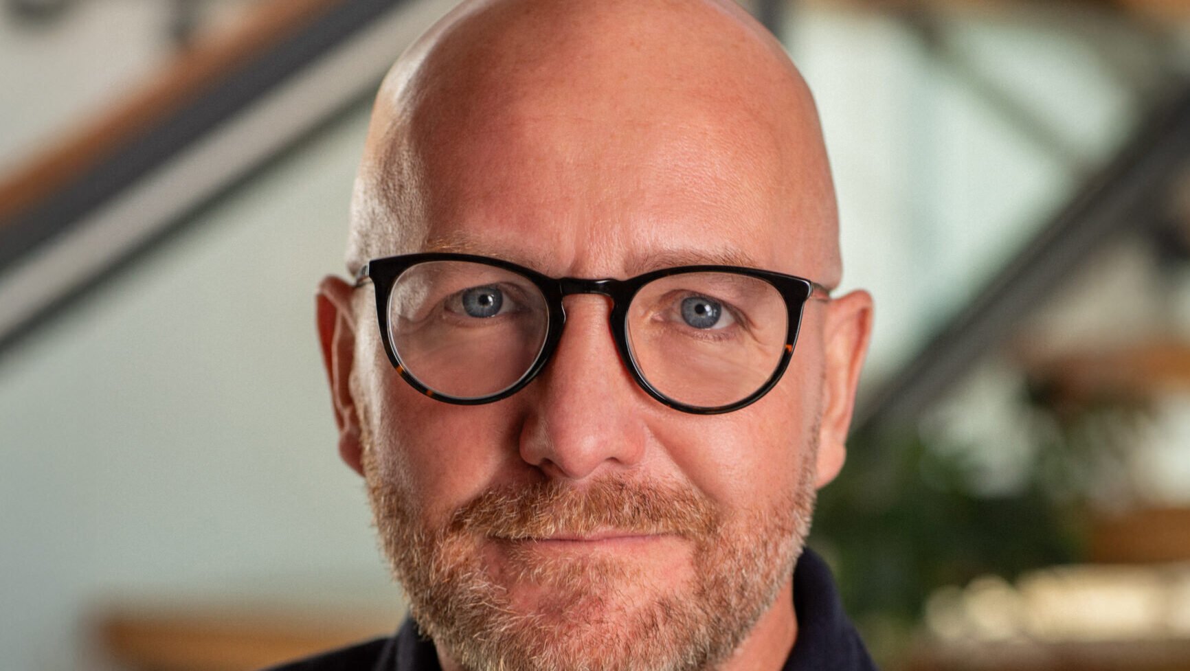 A bald man with glasses and a trimmed beard, wearing a dark buttoned shirt, sits indoors—reminiscent of a creative mind behind viral marketing campaigns—with a blurred staircase in the background.