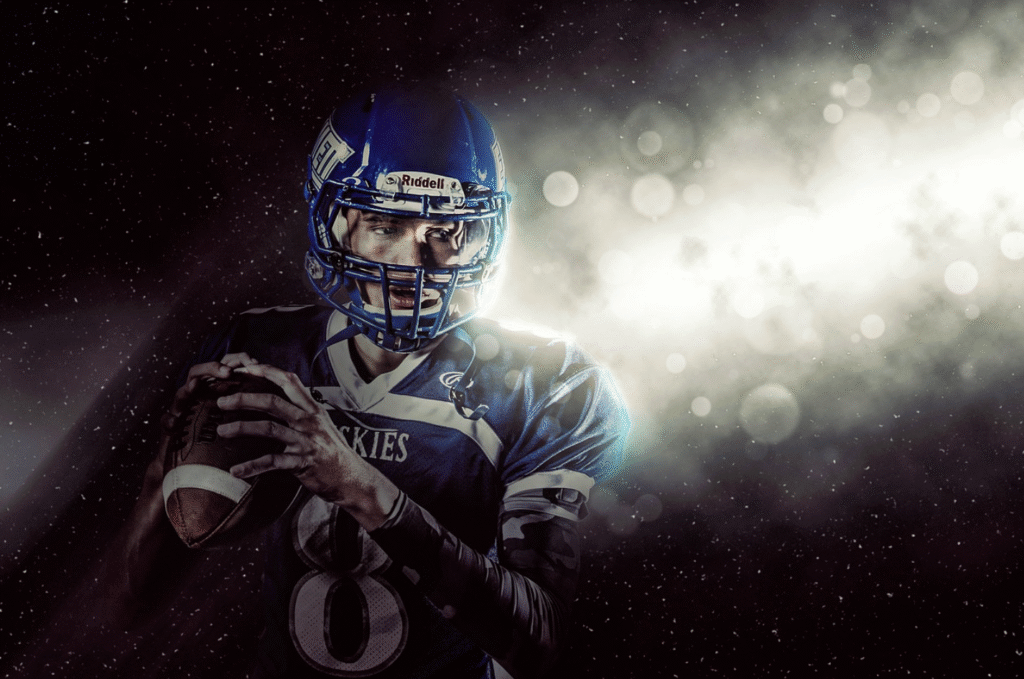 A football player in a blue uniform and helmet holds a football, preparing to throw under dramatic lighting and a smoky background, ready for the Big Game.