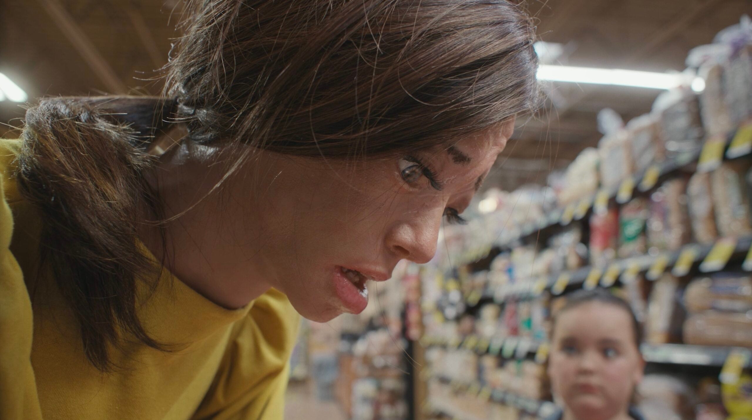 A woman in a yellow shirt looks down with a serious expression in a grocery store aisle, perhaps searching for Hand-Built or Go Raw products, while a child is visible in the blurred background.