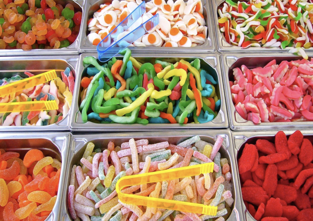 Assorted colorful gummy candies displayed in metal trays, with plastic tongs placed on top for serving—like a scene from cinematic shopping lists where candy-crushed arenas tempt every sweet tooth.