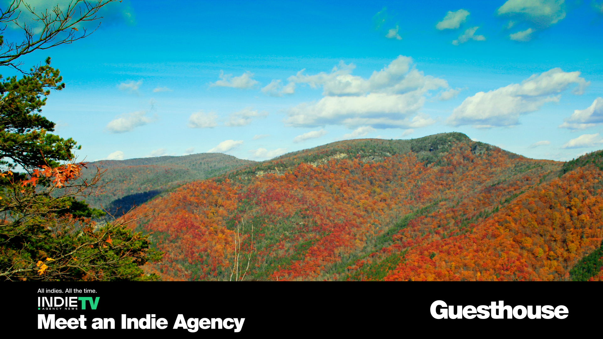 A panoramic view of tree-covered mountains with autumn foliage under a blue sky, overlaid with text promoting Indie TV and Guesthouse.