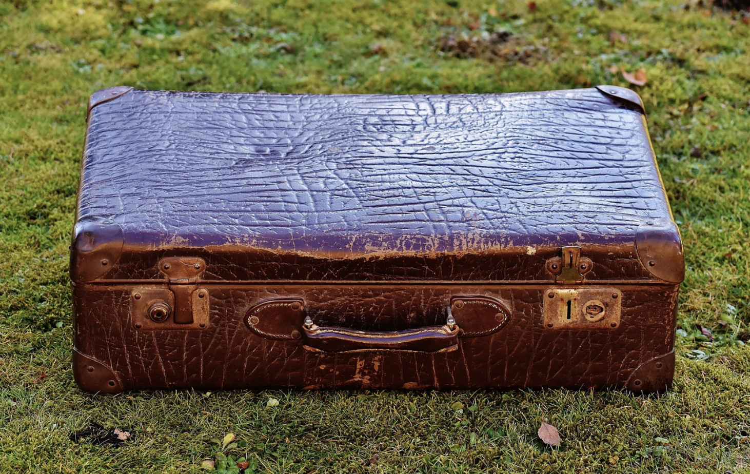 A worn brown vintage suitcase with a textured surface and metal clasps sits on green grass, as if left behind at empty auctions.