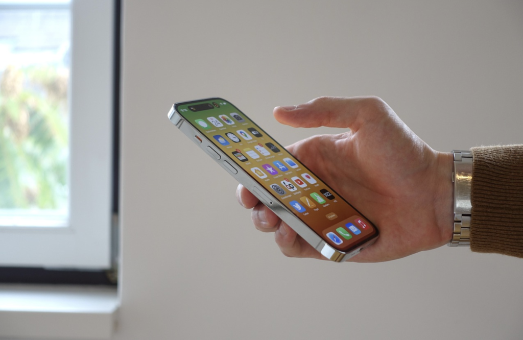 A hand holding a smartphone with the screen displaying several app icons, near a window with natural light.