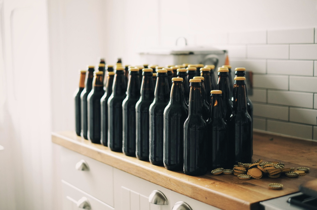 Seventeen dark glass bottles with caps sit on a kitchen countertop, with loose bottle caps scattered nearby.