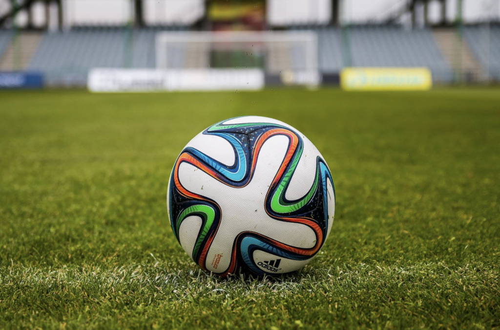 A colorful Adidas soccer ball rests on a green grass field with a goal and empty stadium seats visible in the background, capturing the vibrant energy of the World Cup.