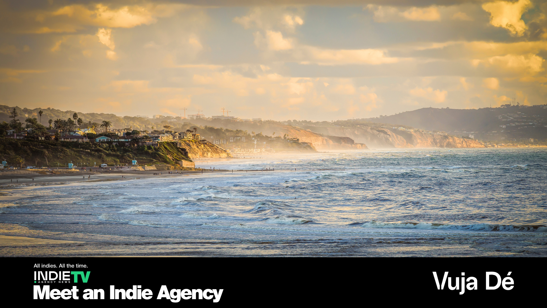Waves roll onto a sandy beach with coastal cliffs and houses under a cloudy sky at sunset. Text reads Meet an Indie Agency and Vuja Dé. Experience the difference with Vuja Dé Digital, a digital agency redefining client communication in every project.