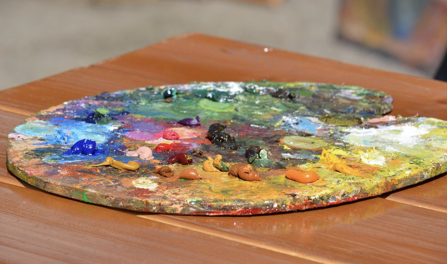 A wooden artists palette with various colorful paint blobs rests on a brown wooden table in bright daylight, reminiscent of the vibrant scenes seen in Presidential Portraits.