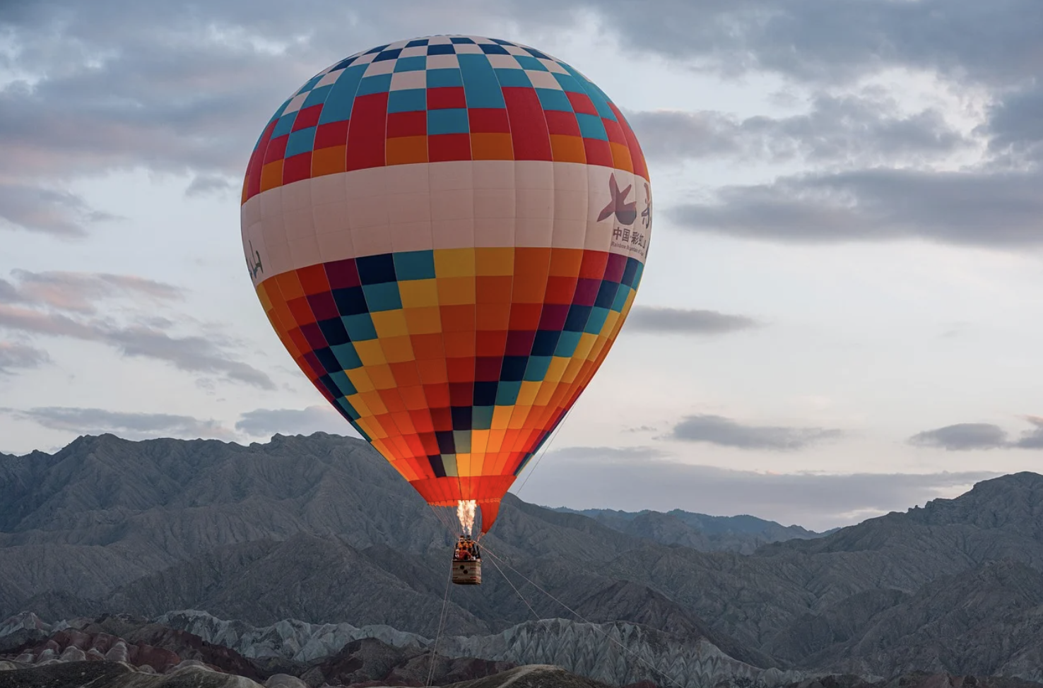 A colorful hot air balloon floats above a mountainous landscape at dusk, with a flame visible beneath the balloons envelope.