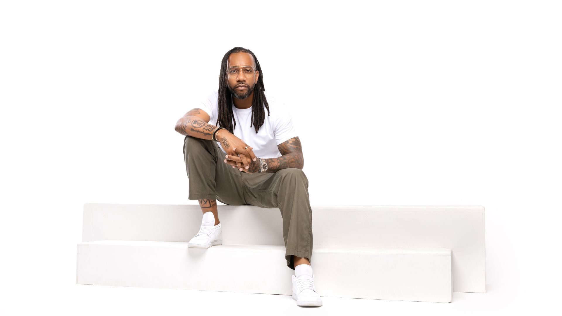 Man with long hair, glasses, and tattoos sits on white steps. He wears a white t-shirt, khaki pants, and white sneakers against a plain white background.