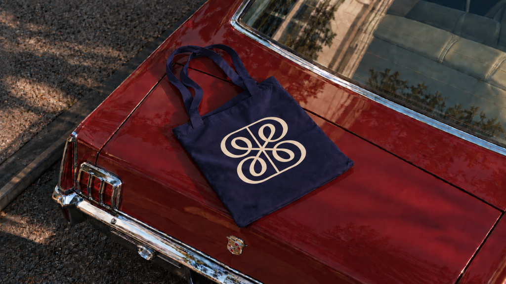 A navy blue tote bag with a white abstract design lies on the trunk of a red vintage car—a perfect shot for a Bensimon Byrne new pitch, with trees reflected on the car’s surface.