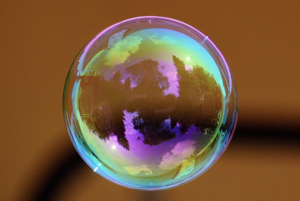 Close-up of a soap bubble reflecting trees, sky, and surrounding environment, with colorful iridescent patterns on its surface against a blurred brown background.