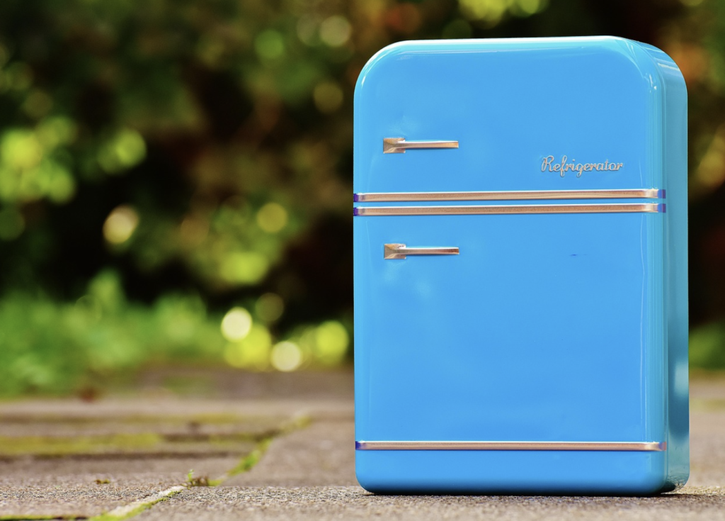 A retro-style blue refrigerator stands outdoors on a stone surface with greenery in the background.