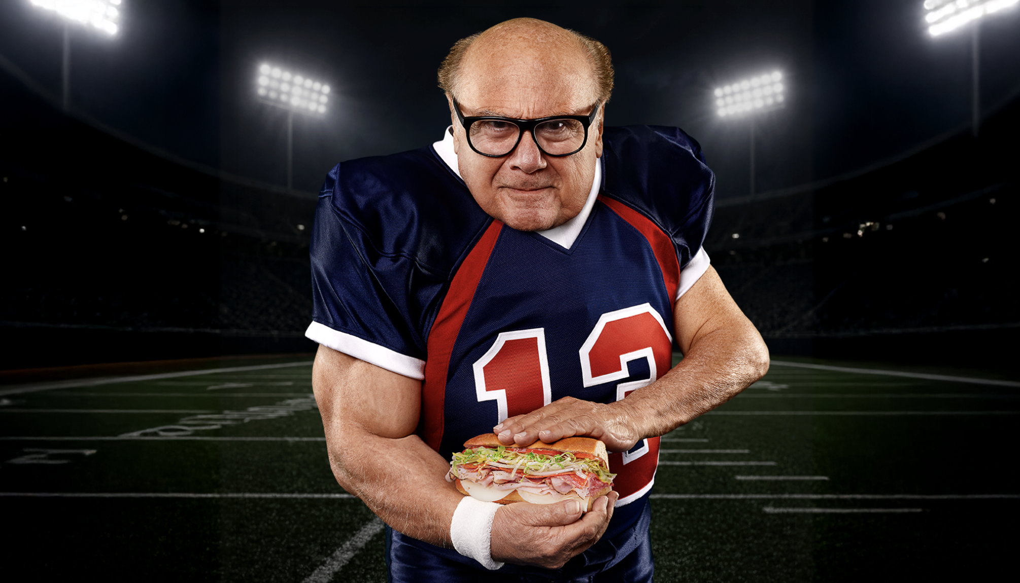 A man in a football uniform with the number 13 stands on a stadium field, holding a large sub sandwich with both hands, reminiscent of Eli Manning celebrating after making a game-winning play.