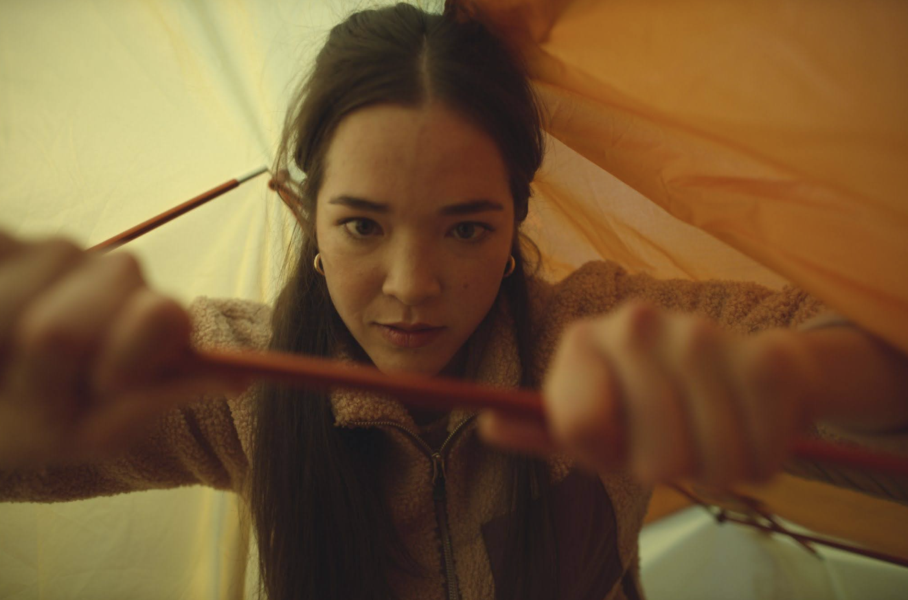 A woman inside a tent is assembling tent poles with both hands, looking focused. The tent fabric is yellow and orange, and sunlight filters through.