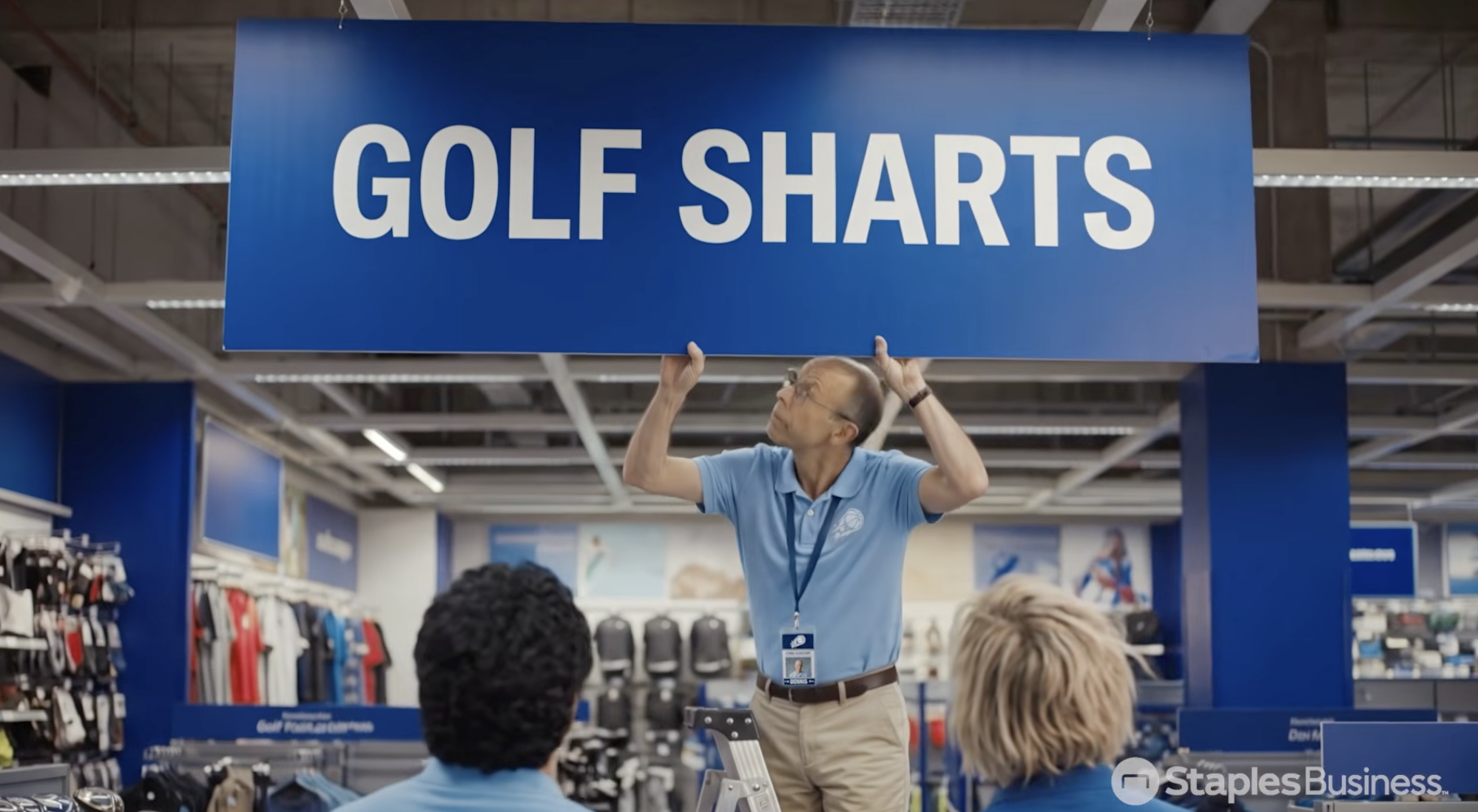 A man adjusts a large blue sign in a store that reads GOLF SHARTS instead of GOLF SHIRTS, while two people look on—perhaps wishing for an easy button from Staples to quickly fix the embarrassing mix-up and avoid turning these shirts into unwanted products.