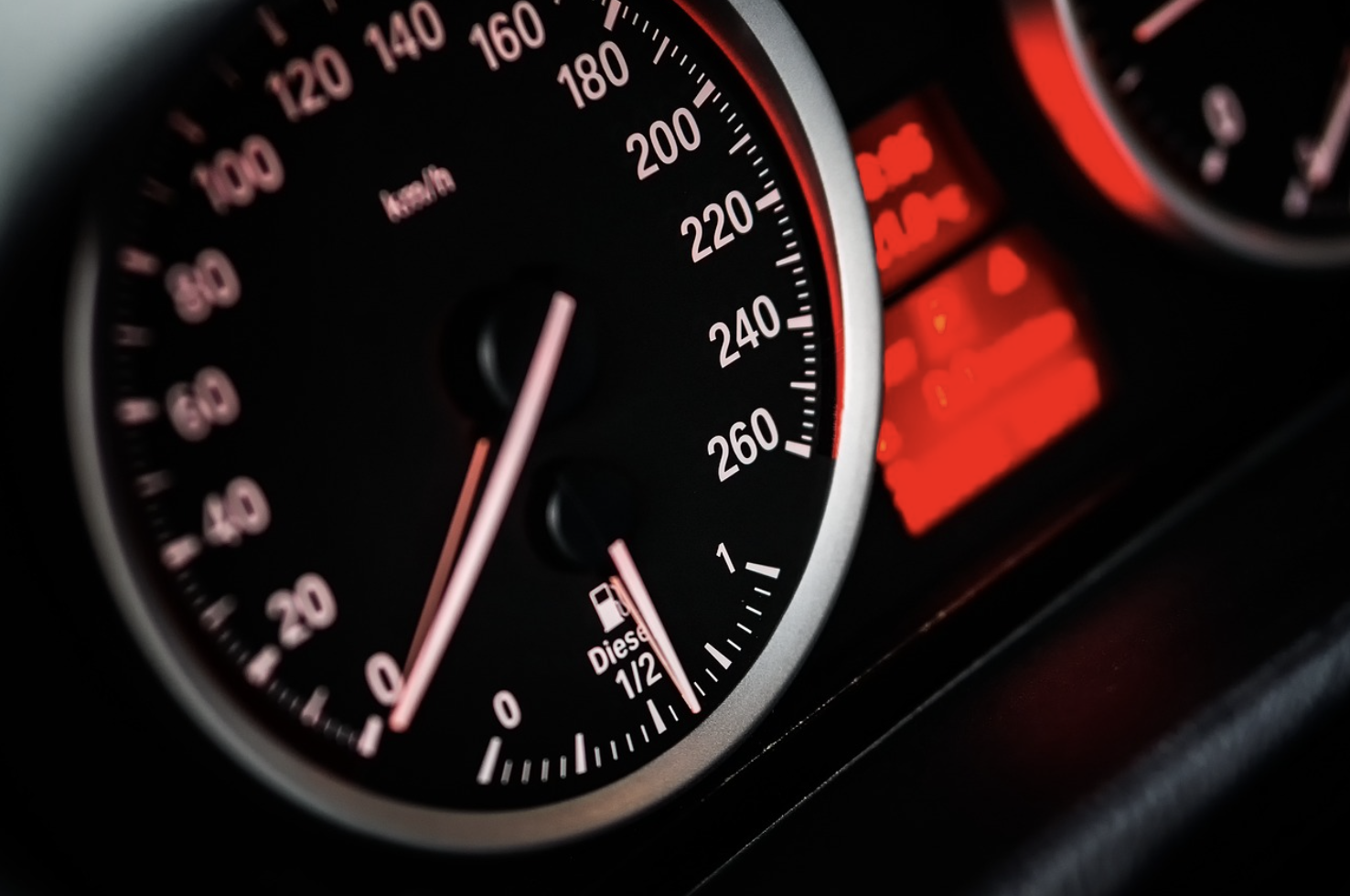 Close-up of a car dashboard speedometer and fuel gauge, showing a speed of 0 km/h and a nearly full diesel tank, with red dashboard lights in the background—capturing the calm before One Airing into busy traffic.