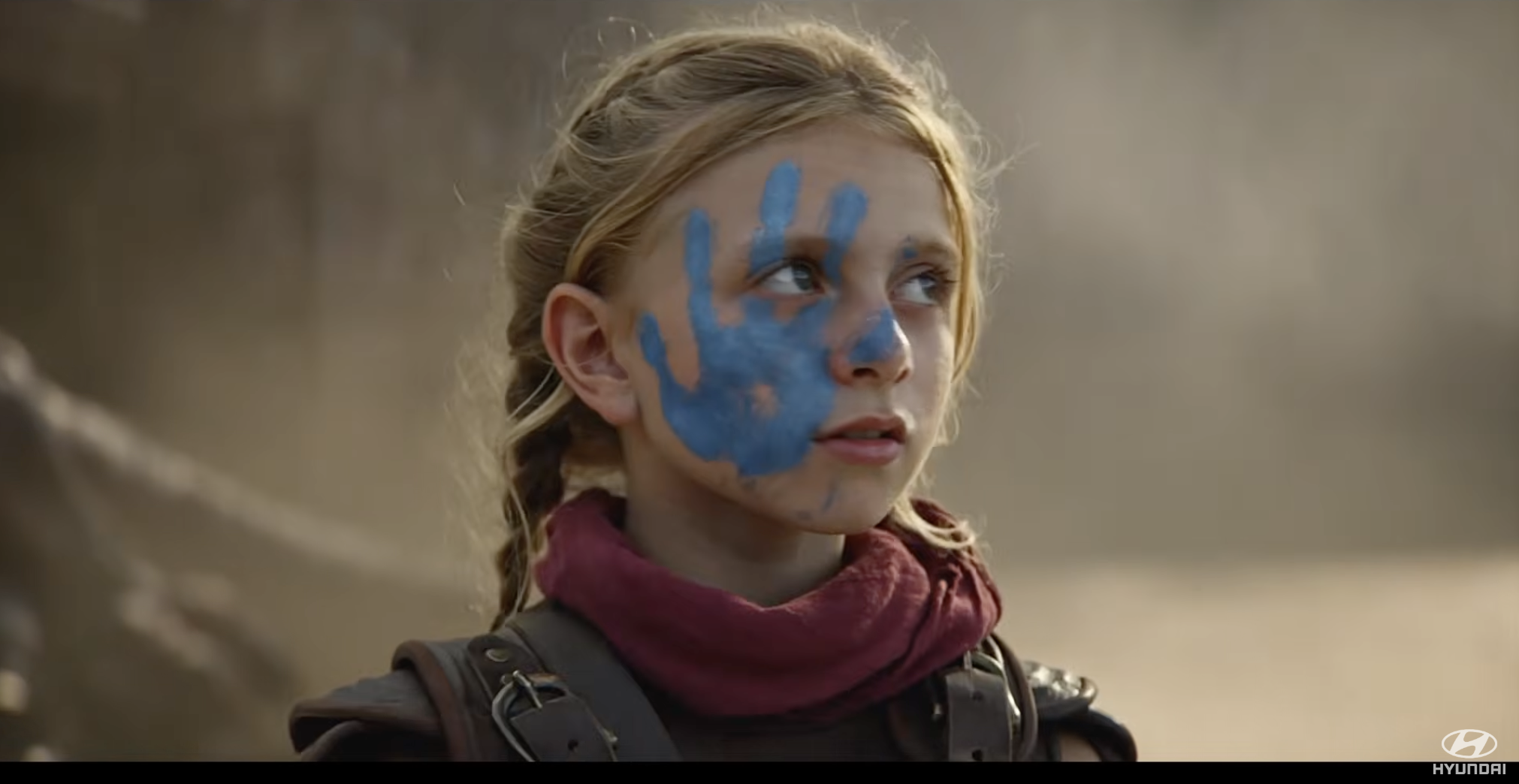 A young girl with a large blue handprint painted on her face looks to the side, wearing a scarf and brown clothing like armor, with a blurred natural background.