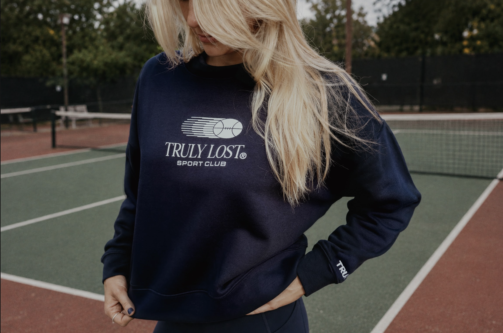 A person with long blonde hair stands on a tennis court wearing a navy blue TRULY LOST Sport Club sweatshirt, looking down with hands near the hem.