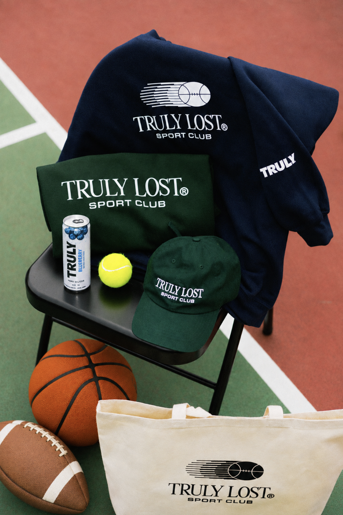 Assorted Truly Lost Sport Club merch for fans is displayed on a chair and court, featuring a tennis ball, can, basketball, football, and tote bag.
