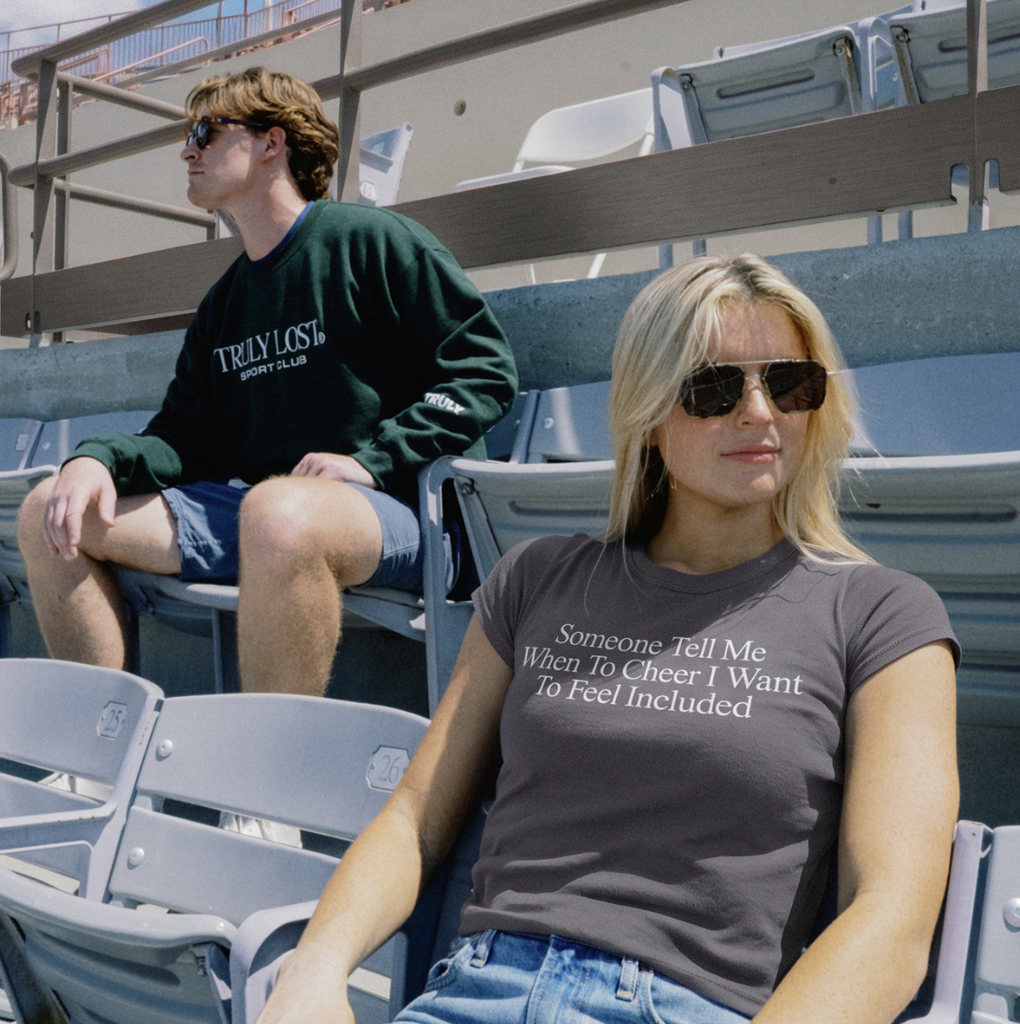 Two fans sit in stadium seats. The woman in front wears sunglasses and a shirt from the Truly Lost merch line that says, Someone Tell Me When To Cheer I Want To Feel Included. The man behind her looks to the side.