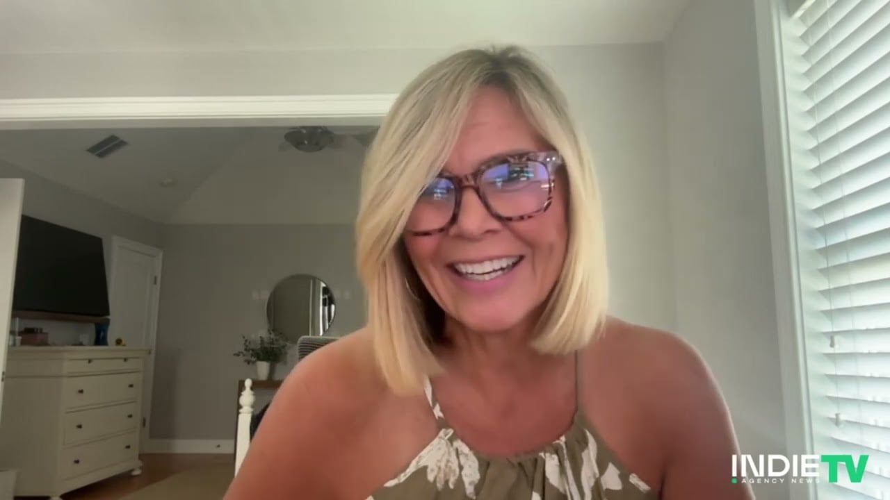 A woman with blonde hair and glasses smiles while seated indoors near a window, celebrating 20 years of innovation with Logitech, with INDIE TV branding in the bottom right corner.