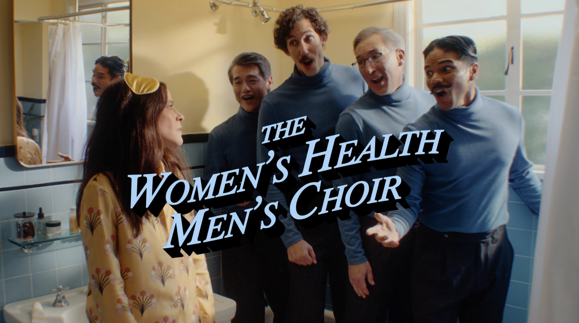 A woman stands in a bathroom in pajamas and a sleep mask while four men in blue shirts sing to her. Text on the image reads, The Womens Health Mens Choir.