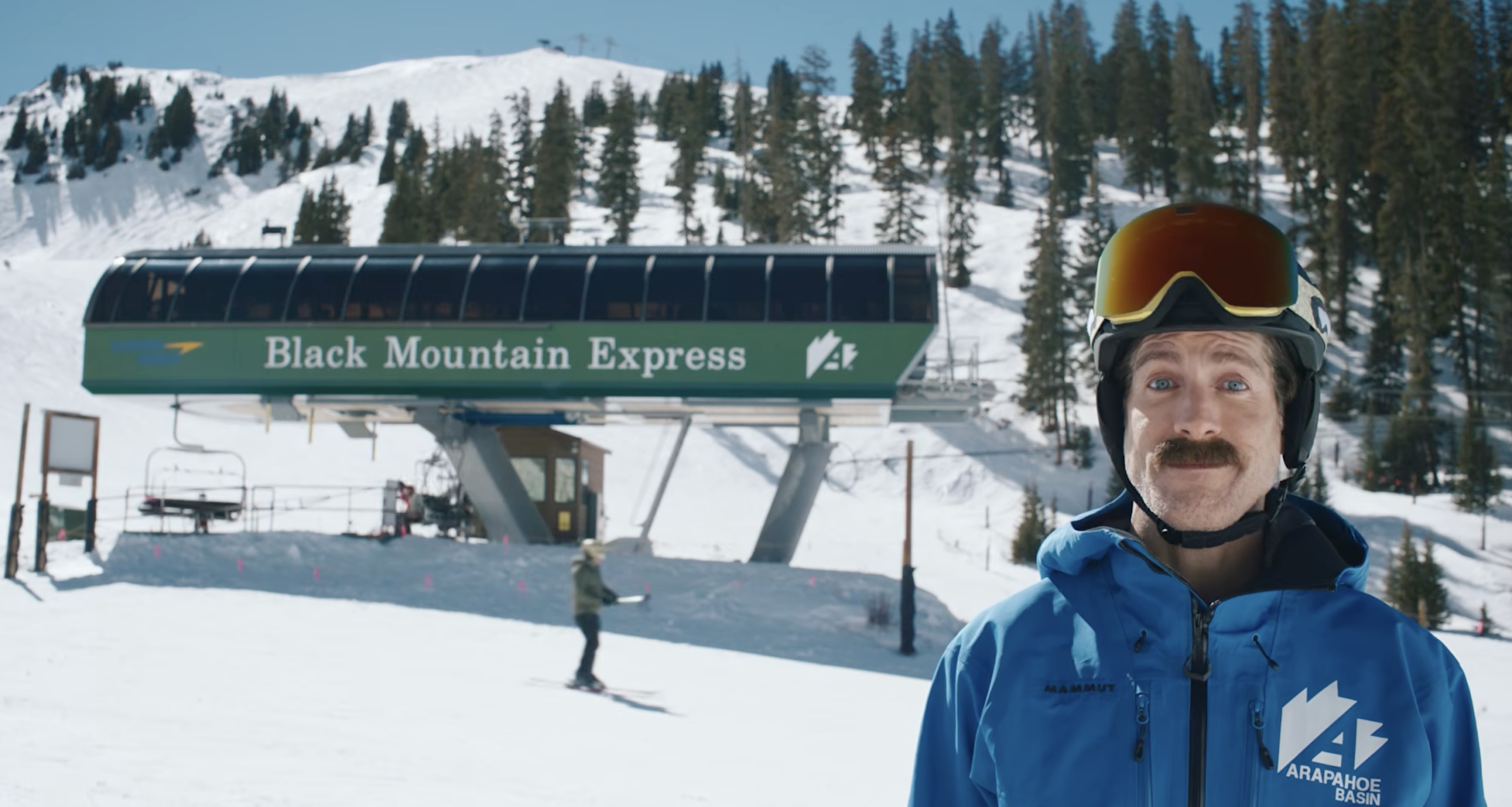 A mustachioed man wearing a ski helmet and blue jacket stands in front of the Black Mountain Express chairlift at Arapahoe Basin for a day of skiing.